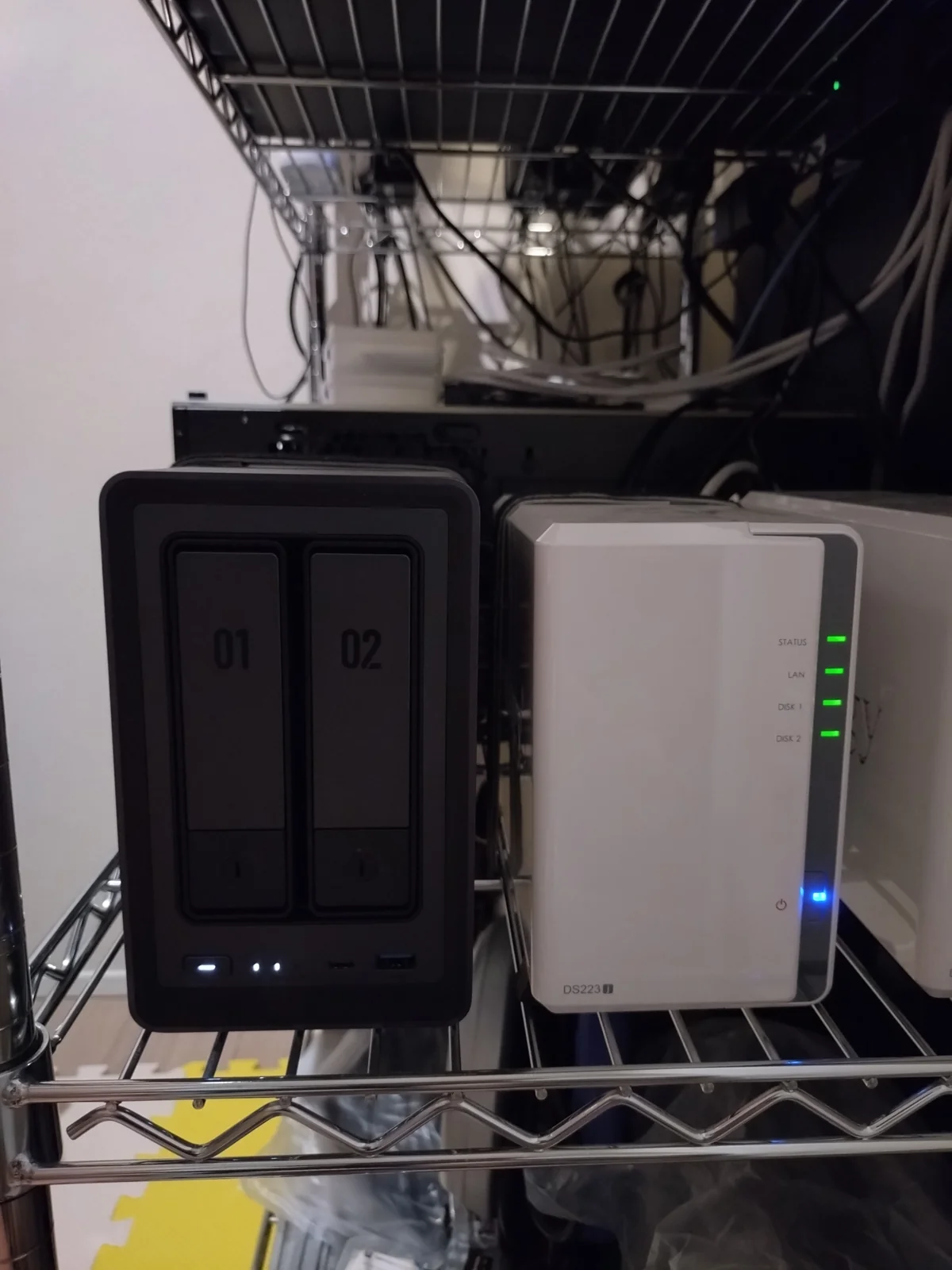 Synology DS223j(右・白・STATUS/LAN/DISK1/DISK2の4個LEDが緑点灯・電源LEDも青点灯)を筆者宅のメタルラックで24時間稼働中の実機写真。左には比較用のUGREEN DXP2800も並んでいる