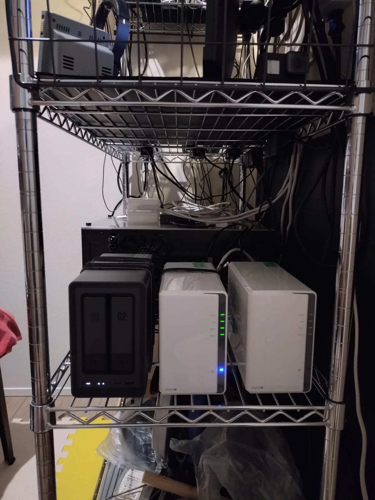 納戸2帖のアルミラックに設置された3台のNAS：手前がUGREEN DXP2800（黒・稼働中）、中央がSynology DS223j（白・LED4個緑点灯・稼働中）、奥が前世代のSynology DS216j（白・引退置物）
