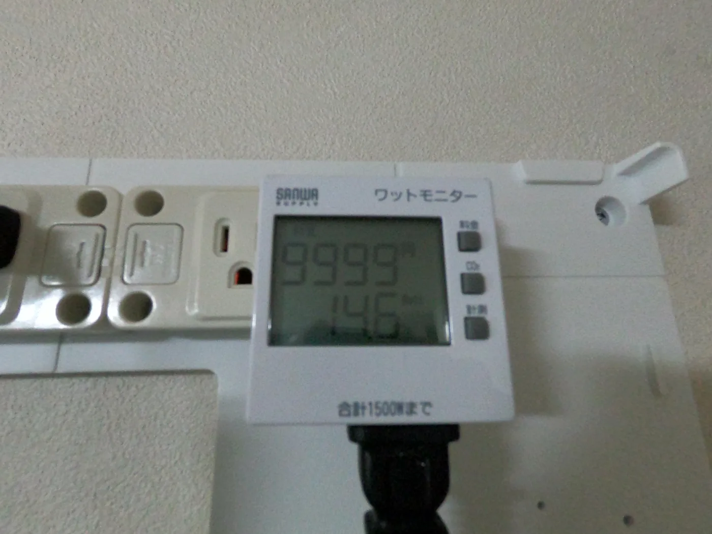 サンワサプライのワットモニターの液晶表示。上段に9999、下段に14.6Wの数値が出ており、壁付けの電源タップに接続されている